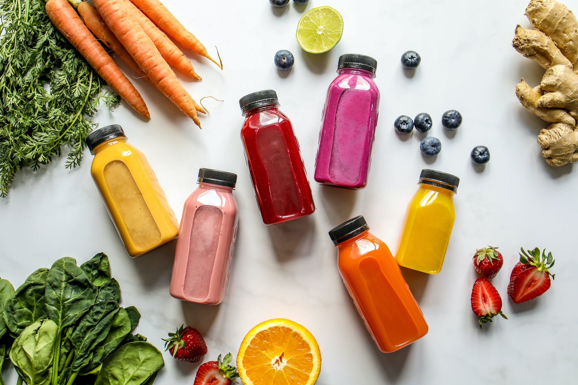 colorful bottles with smoothies beside carrots ginger leaves and berries